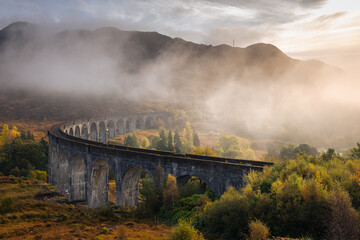 Fototapeta premium The morning train steams across the Glenfinnan Viaduct, its plume rising against the Highland mist. Golden light touches the hills, creating a scene of timeless beauty in Scotland’s heart.