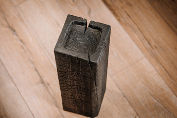 Charred square wood block standing upright on a wooden floor, with visible cracks and rings on the...