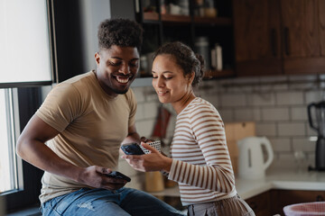 A happy couple enjoys a casual moment in their kitchen, looking at a smartphone together while sharing laughter. A breakfast spread with fruit and pastries is visible on the counter.