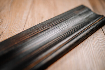 Close-up of a dark wooden molding plank with a weathered texture, placed diagonally on a light wood...