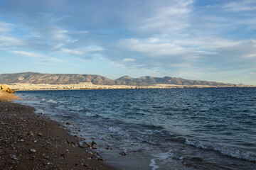 Sun illuminates the urban outskirts of Athens on the shores of the gulf. Waves lapping at the pebbly beach.