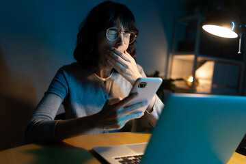 Tired woman yawning in front of laptop, working late at night using phone.