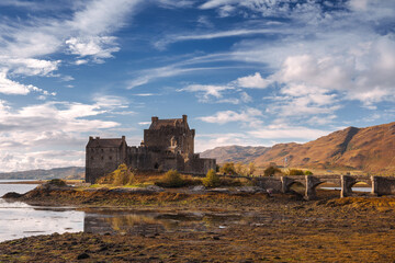 Fototapeta premium Eilean Donan Castle stands majestically amid autumn hues, its reflection shimmering in calm waters. Misty Highlands and golden trees create a scene of timeless beauty.