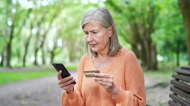 Concerned mature woman sitting outdoors holding phone and credit card, reflecting on online scams and risks of fraud in digital transactions. Elderly female with gray hair became a victim of deception