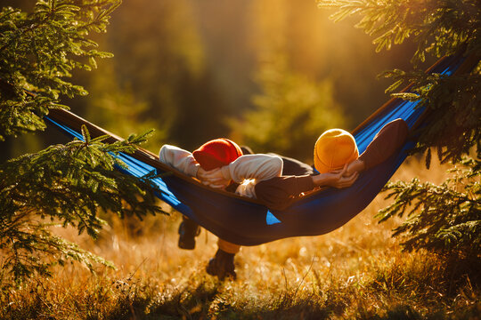 Couple of tourists resting in a nylon hammock hanging between fir trees, mountain landscape background at sunset. Family camping in the mountains.