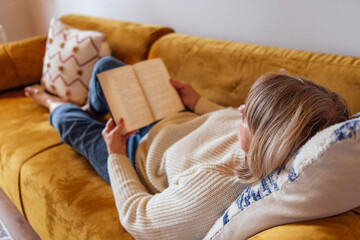 An elderly woman in casual clothes is lying on the couch and reading a novel. A mature lady holds a...