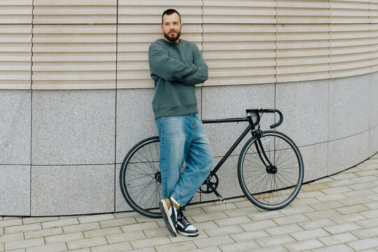 A man in a casual outfit stands confidently with his arms crossed next to a black bicycle. He is in an urban area.