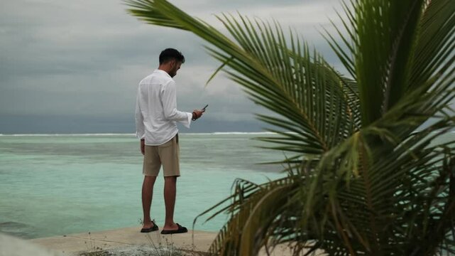 A young man in light clothes with phones in his hands stands on a sandy beach under palm trees. Against the background of the waves of the sea and a cloudy sky. Maldives.