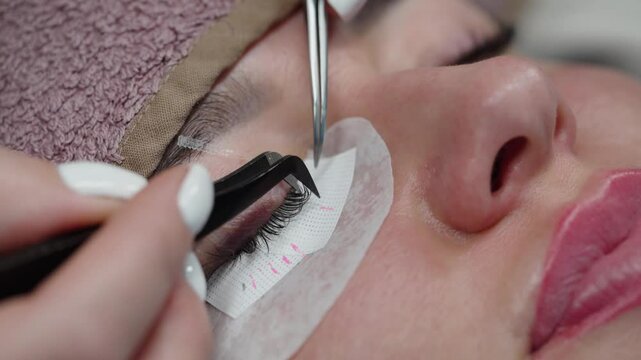 Close-up view of a beautician applying eyelash extensions with tweezers to a client&rsquo;s eye during a professional lash treatment. Commercial use.
