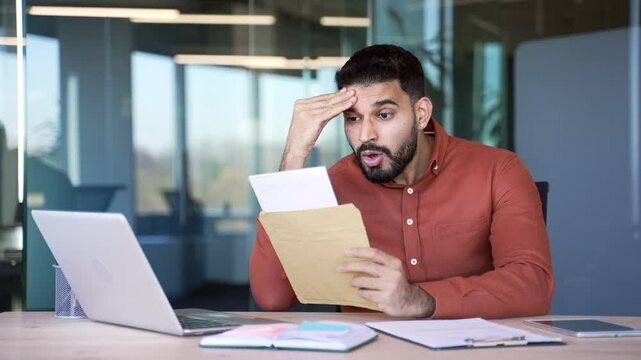 Upset disappointed businessman reading a letter with bad news sitting in a business office. Worried entrepreneur frustrated by receiving unpleasant negative notification after opening the envelope