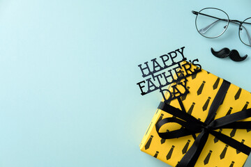Happy Father's Day flat lay: Gift box, greeting sign, eyeglasses, and mustache prop on a blue background. Top view with copy space for celebration concepts.