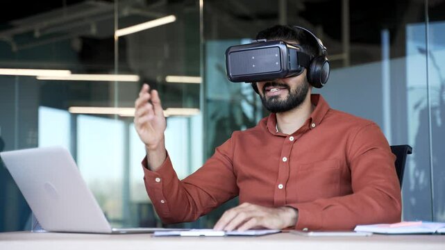 Successful businessman works using VR glasses in virtual reality simulator sitting at workplace in business office. Worker uses gestures to control computer programs and flips through virtual pages