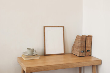 Scandinavian home interior. Cup of coffee tea, old books on table. White wall background. Empty wooden vertical picture frame mockup. Wicker magazine file folder holder, boho rattan organizer.