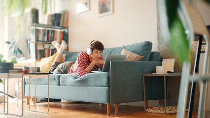 Side view of Caucasian woman in white headphones laying on soft couch. Enjoying weekend in cosy...