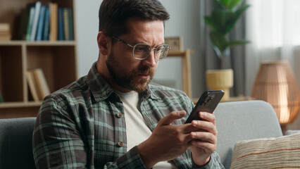 Caucasian adult man male guy at home living room nervous looking at mobile phone bad news amazed shocked close mouth with hand message from bank stressed smartphone notification online error scam