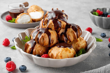 Profiteroles with vanilla cream, fresh berries and chocolate glazed in a bowl on a gray concrete background. A delicious French dessert.