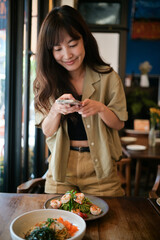 Asian woman joyfully captures photo of pasta dish with phone, sharing experience on social media. vibrant meal and cozy restaurant setting create warm and inviting atmosphere