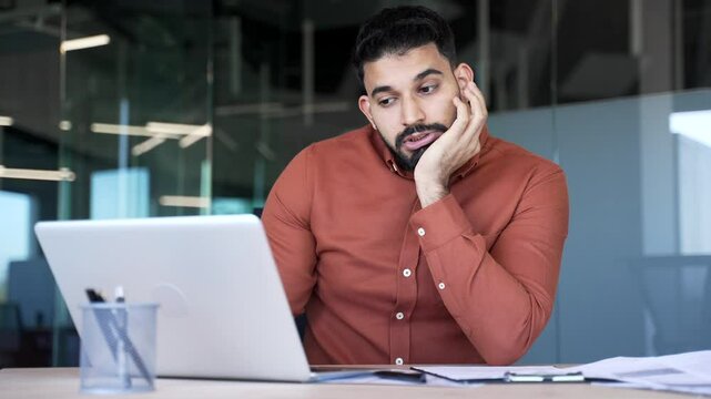 Exhausted businessman is bored sitting at workplace at desk in business office. Upset bearded man is tired from working with laptop and doing tasks. A lazy worker procrastinates, gets distracted