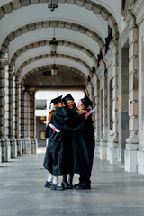 Group of graduated students wearing gowns and holding diplomas are hugging under university archway