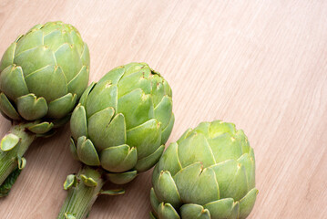 Obraz premium Three vibrant green artichokes are aligned against a natural woodgrain surface, showcasing their fresh and symmetrical appeal for culinary or artistic contexts. Cooking ingredients, healthy food