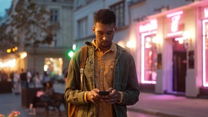 Portrait of focused relaxed young African American male tourist communicating online texting with phone standing in city center with beautiful neon signs on background. Evening concept.