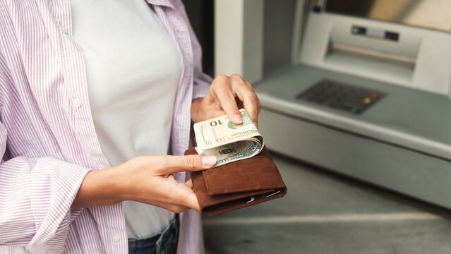 Extreme close-up view of successful caucasian businesswoman withdrawing money from credit card using ATM. Young adult female cashing out dollars bills putting in wallet. Finance concept.