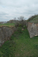 Aerial view of the lower ramparts of Petrovaradin Fortress in Novi Sad, Serbia. Overgrown grass, stone walls, and cloudy skies highlight historical architecture and peaceful decay.