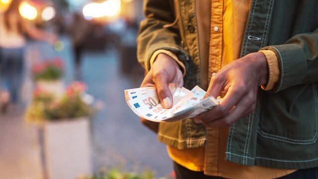 Close-up view of young African-American man counting euro notes putting it in pocket. Adult successful male holding salary income planning budget standing on street in evening. Finance concept.