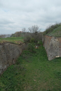 Aerial view of the lower ramparts of Petrovaradin Fortress in Novi Sad, Serbia. Overgrown grass, stone walls, and cloudy skies highlight historical architecture and peaceful decay.