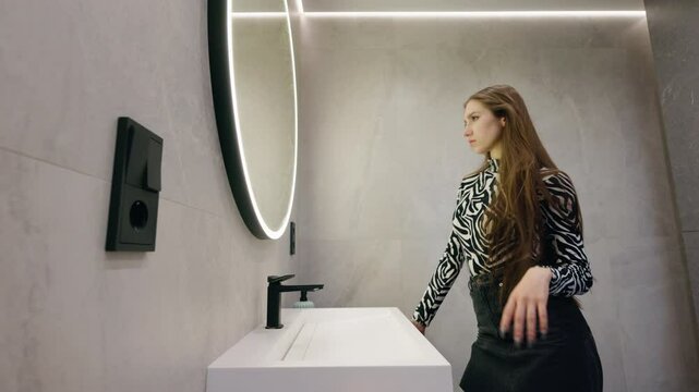 Young woman adjusting her long hair while standing by a modern sink and round mirror in a stylish bathroom