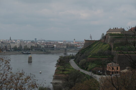 Panoramic view of Novi Sad, Serbia, with the Petrovaradin Fortress on the right, the Danube River, and the city skyline in the background. Overcast spring day with visible urban and historic landmarks