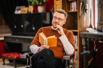 Fototapeta premium A man with glasses and a beard reads a book while seated in a cozy room. He appears calm and focused, holding his chin thoughtfully..