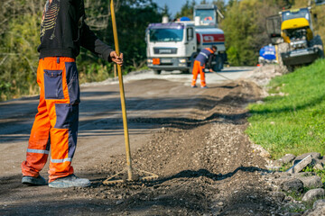 Worker holding a rake and another spreading gravel in the background, smoothing the road base for asphalt on a countryside road construction site.