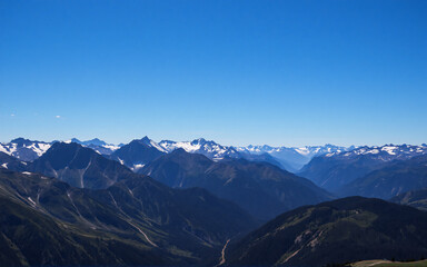 A wide view of a mountain range stretches under a cloudless blue sky. Some peaks are covered in snow, contrasting with the dark green slopes below in this scenic landscape.