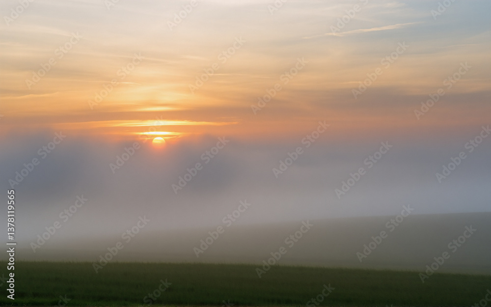 Poster A tranquil scene of sunrise or sunset with warm light filtering through a thick fog blanketing a field. The sun subtly illuminates the hazy atmosphere and soft clouds above.