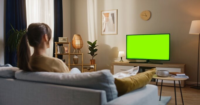 Camera sliding behind blurred Caucasian woman with ponytail. Holding focus on TV screen with chroma key. Female sitting on couch and switching something once with TV remote. Resting in modern room.