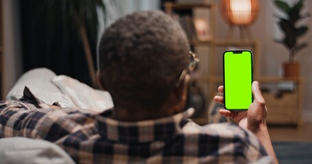 African American man lying at sofa and holding chroma key phone. Man having video chat lying at couch. Male relaxing at comfortable home. Man holding smartphone with green template gadget.