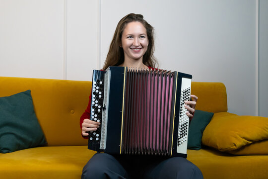 Happy musician playing russian bayan accordion and smiling while relaxing on a cozy yellow sofa in her living room, fully immersed in her beloved passion for music and creativity