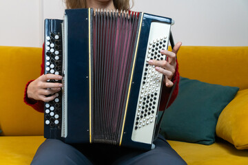 Woman musician wearing a red sweater sits comfortably on a yellow sofa in a cozy living room,...