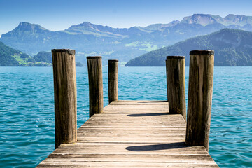 Fototapeta premium Jetty with rustic poles above blue green water of the lake lucerne. Mountains and hills in the background.