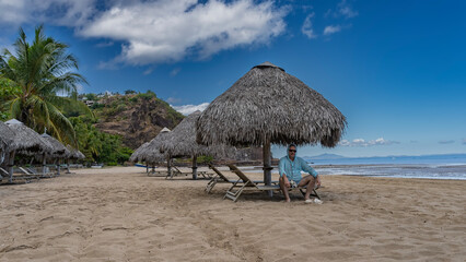 A row of deck chairs under straw umbrellas on a sandy beach by the ocean. A man in sunglasses is sitting on a sunbed, smiling. Flip-flops are nearby. A hill in the distance.Palm tree, blue sky, clouds