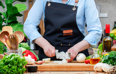 Housewife cuts onion into half in cozy kitchen with wooden table, kitchenware, vegetables, herbs and ingredients for cooking. Healthy vegan food, culinary, recipes, blogging