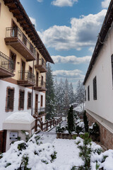 Traditional Alpine buildings with wooden balconies and sloped roofs stand amidst snow covered ground, trees, and a cloudy winter sky in Cortina, Italy.