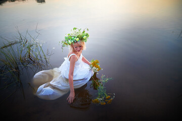 A girl in a white sundress, a wreath of flowers and a bouquet in the water in the evening at...