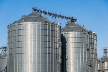 Large steel silos dominate the landscape of an agricultural site, surrounded by equipment and machinery against a vibrant blue sky at noon