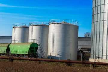 Three tall metallic silos stand prominently alongside a green train car in an industrial area, with bright blue sky and minimal clouds above