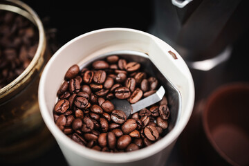 Roasted coffee beans in an electric coffee grinder on a table with a moka pot and cup, coffee preparation