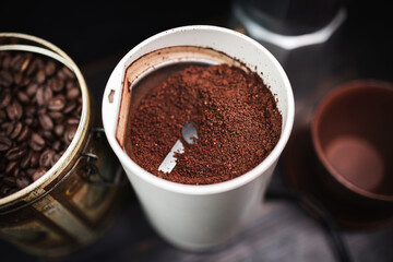 Ground coffee in an electric coffee grinder on a wooden table with a moka pot and a cup, coffee preparation