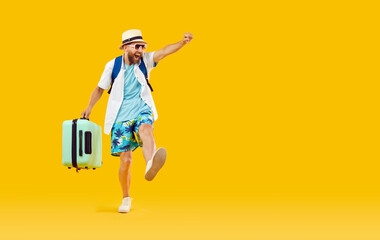 Happy, funny, excited man tourist in summer shirt, shorts, hat and glasses carrying his suitcase and walking in on orange yellow color studio background. Holiday, vacation, travel, fun, trip concept