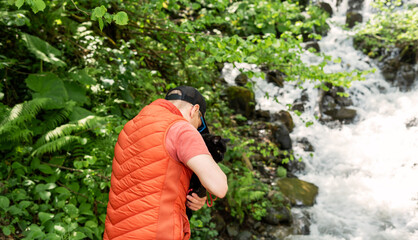 Man wearing an orange vest holds a black cat next to a cascading waterfall in lush forest....